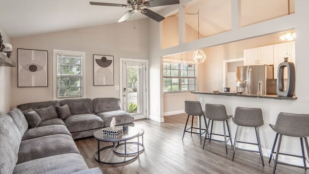 A spacious living room with a large gray sectional sofa, a round coffee table, and a ceiling fan. There are three bar stools and a kitchen area visible through an open door.