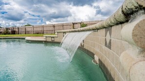 A swimming pool with a waterfall feature, surrounded by a wooden fence and a cloudy sky.