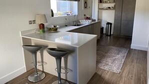 Ett modernt kök med en central köksö, två barstolar, ett fönster med en mönstrad gardinkappa och ett rostfritt kylskåp i bakgrunden.