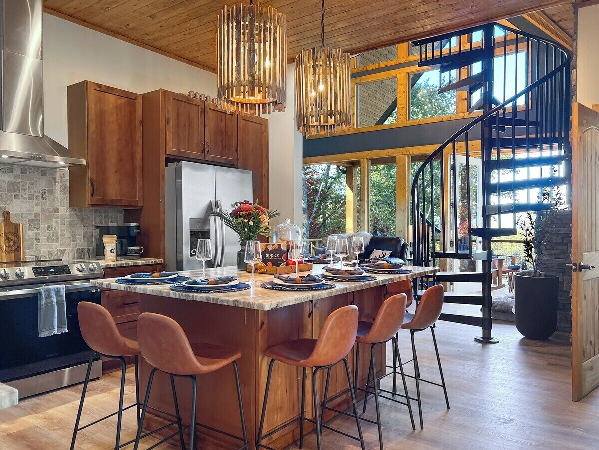 A modern kitchen with a large island, wooden cabinets, and a black staircase leading to an upper level.