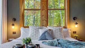 Ein Schlafzimmer mit einem großen Bett, zwei Nachttischlampen, einem Fenster mit Blick auf Bäume und einem Nachttisch mit Pflanze und Tablett.