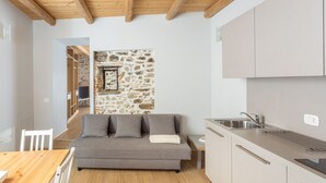 A compact living space with a stone wall, a sofa, a wooden table, and a kitchenette. - La Posteria Junior Suite 1 by icasamia.it (Orino)