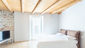 A bedroom with a stone wall, a wooden ceiling, a bed with white bedding, and a television on a stand. - La Posteria Junior Suite 1 by icasamia.it (Orino)