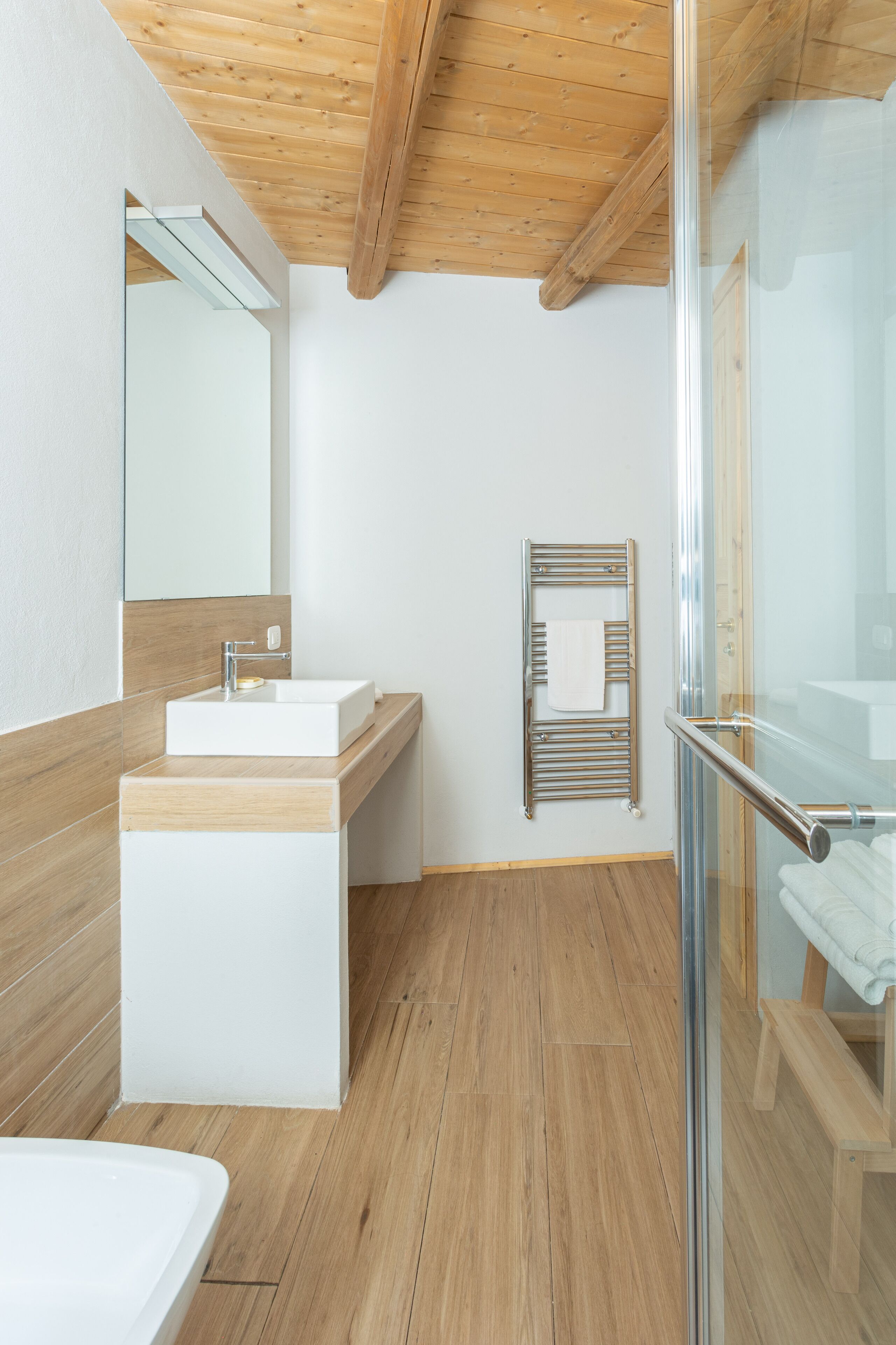 Ein modernes Badezimmer mit Holzboden, einer Duschkabine aus Glas, einem weißen Waschbecken und einem Handtuchhalter.