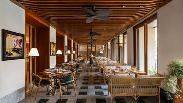 A hotel corridor with dining tables, chairs, ceiling fans, and large windows.