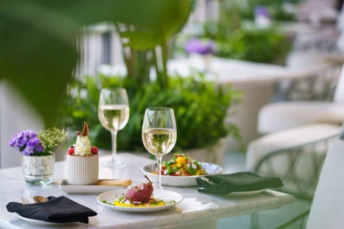 Una mesa de comedor con platos de comida, copas de vino blanco y un pequeño jarrón con flores.