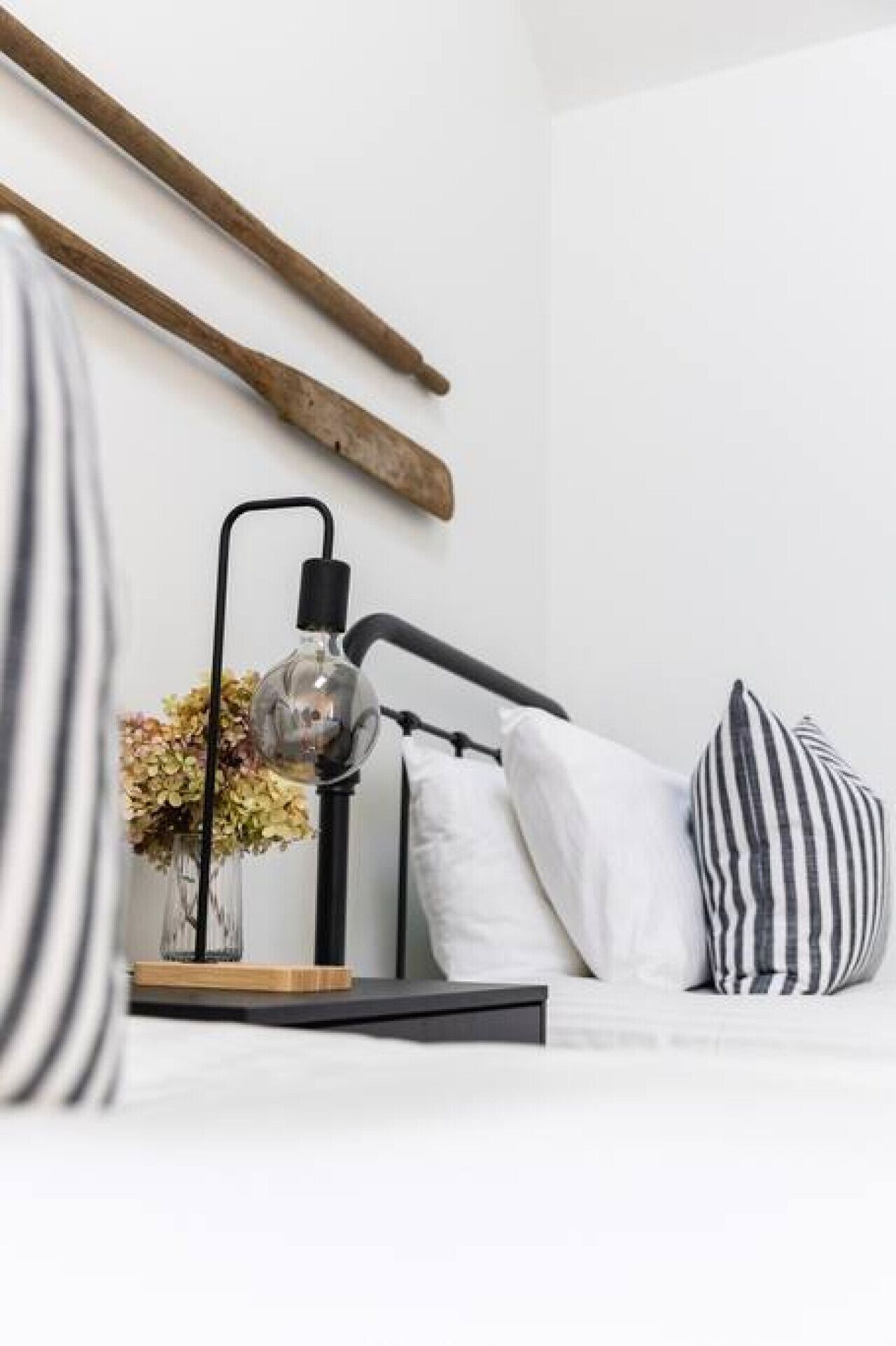 Ein Bett mit schwarzem Metallgestell, ein Nachttisch mit Lampe, eine Vase mit Blumen und gestreifte Kissen.