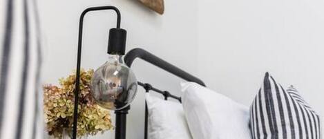 Ein Bett mit schwarzem Metallgestell, ein Nachttisch mit Lampe, eine Vase mit Blumen und gestreifte Kissen.