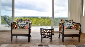 A balcony with two wicker chairs, a small table, and a view of a lush landscape and distant structures. - Cove Bay Villa II "THE PENTHOUSE"  (Anguilla)