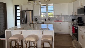 A modern kitchen with white cabinetry, a large island with bar stools, stainless steel appliances, and a view of the outdoors through a window. - Cove Bay Villa II "THE PENTHOUSE"  (Anguilla)