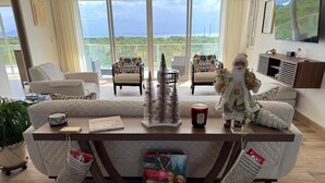A modern living room with a large window, a sofa, a coffee table, and a decorative tree. - Cove Bay Villa II "THE PENTHOUSE"  (Anguilla)