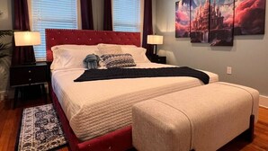 Un dormitorio con una cama grande, dos mesitas de noche, una mesita auxiliar, una ventana con cortinas y una obra de arte colgada en la pared.