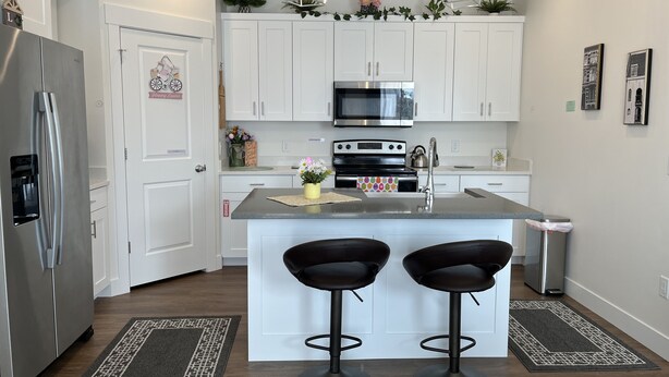 A modern kitchen with white cabinets, stainless steel appliances, and a central island with two bar stools. - Cozy, Modern home in Lindon. (North)