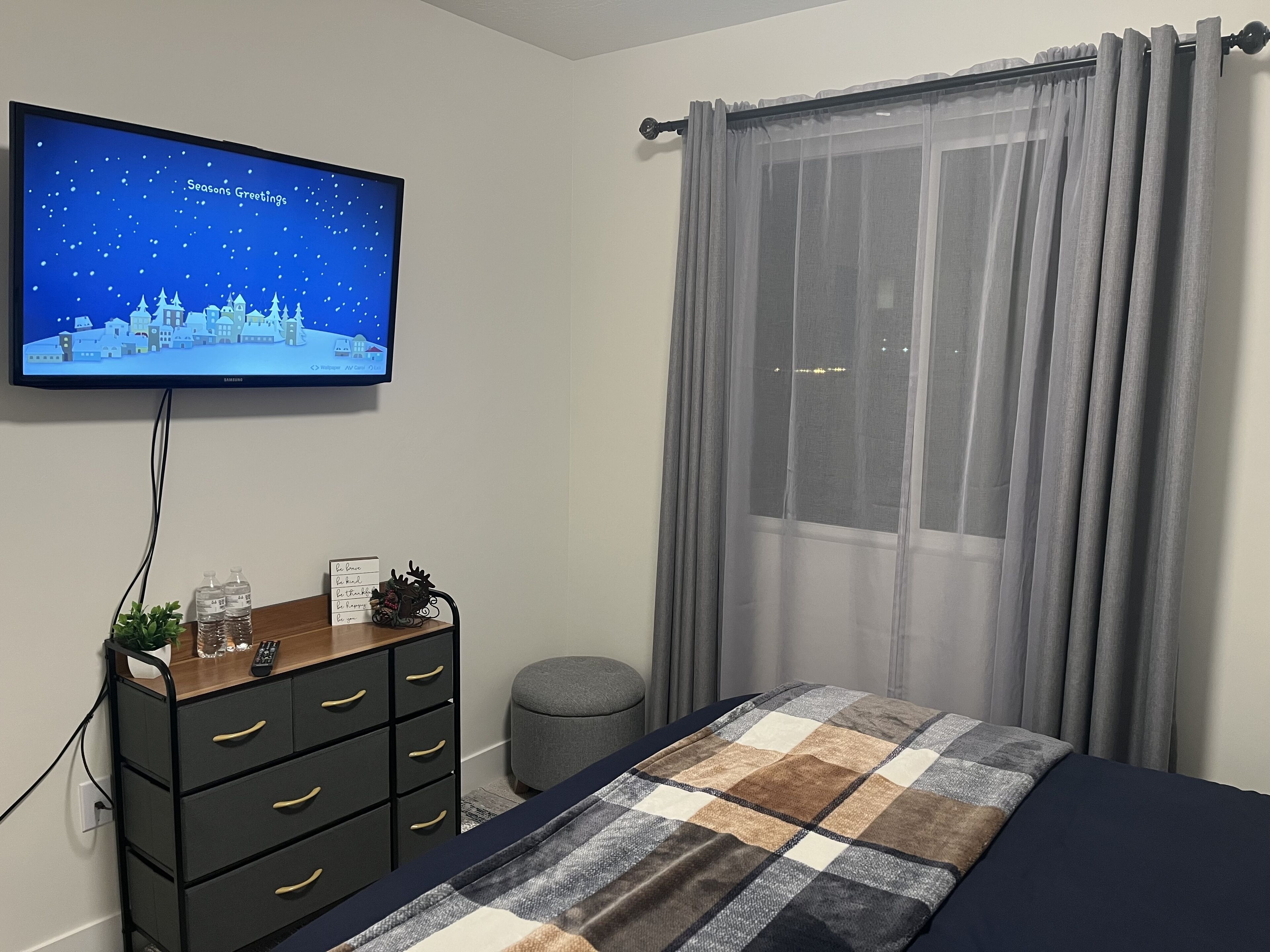 Ein Schlafzimmer mit einem Bett, einem Fernseher, der eine Winterlandschaft zeigt, einem Schminktisch, einem kleinen Hocker und einem Fenster mit durchsichtigen Vorhängen.