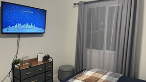 A bedroom with a bed, a TV displaying a winter scene, a dresser, a small stool, and a window with sheer curtains. - Cozy, Modern home in Lindon. (North)