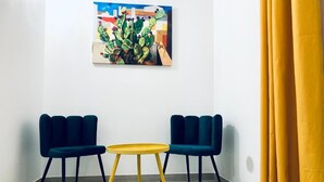 Quarto com duas cadeiras azuis, uma mesa amarela e um quadro de um cacto na parede.