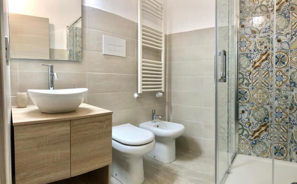 A modern bathroom with a large white sink, two toilets, a shower with a glass door, and a tiled wall with a decorative pattern.