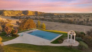 Uma área de piscina com um gazebo, rodeada por jardins bem cuidados e vista para colinas ondulantes.
