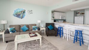 Ein Wohnzimmer mit Sofa, Couchtisch und blauen Barhocker. Es gibt eine Küchenzeile mit weißen Schränken und einem Kühlschrank.