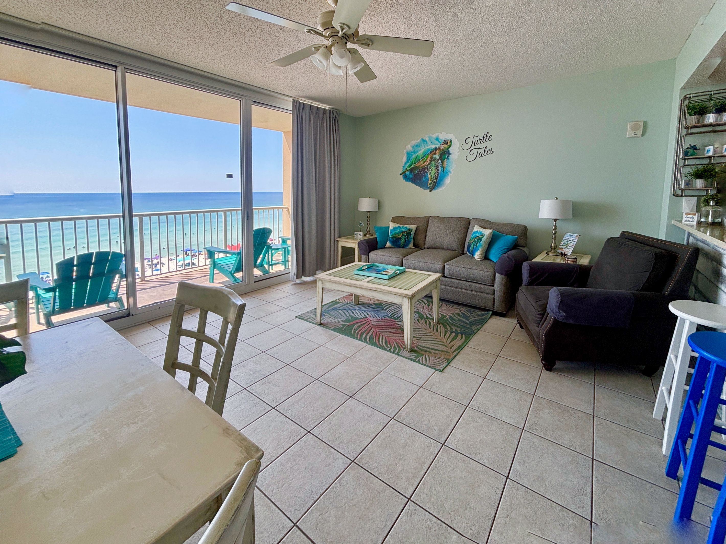 A living room with a view of the ocean, a ceiling fan, a sofa with cushions, a coffee table, and a dining area with chairs.