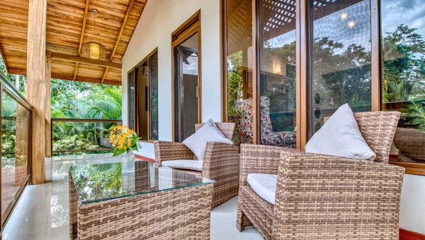 A covered outdoor seating area with wicker furniture and a glass table, surrounded by large windows offering views of greenery.