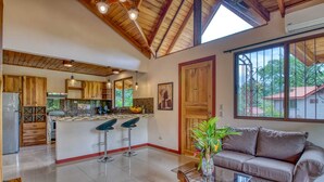 A spacious kitchen with a central island and bar stools, a living area with a sofa and coffee table, and large windows offering a view of the outdoors.