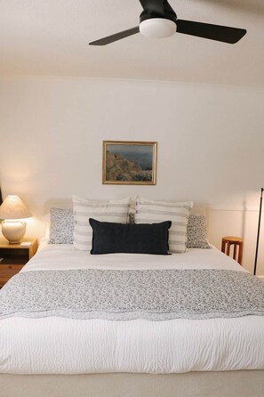 Une chambre à coucher avec un lit, des oreillers, une table de chevet, une lampe, un tableau encadré et un ventilateur de plafond.