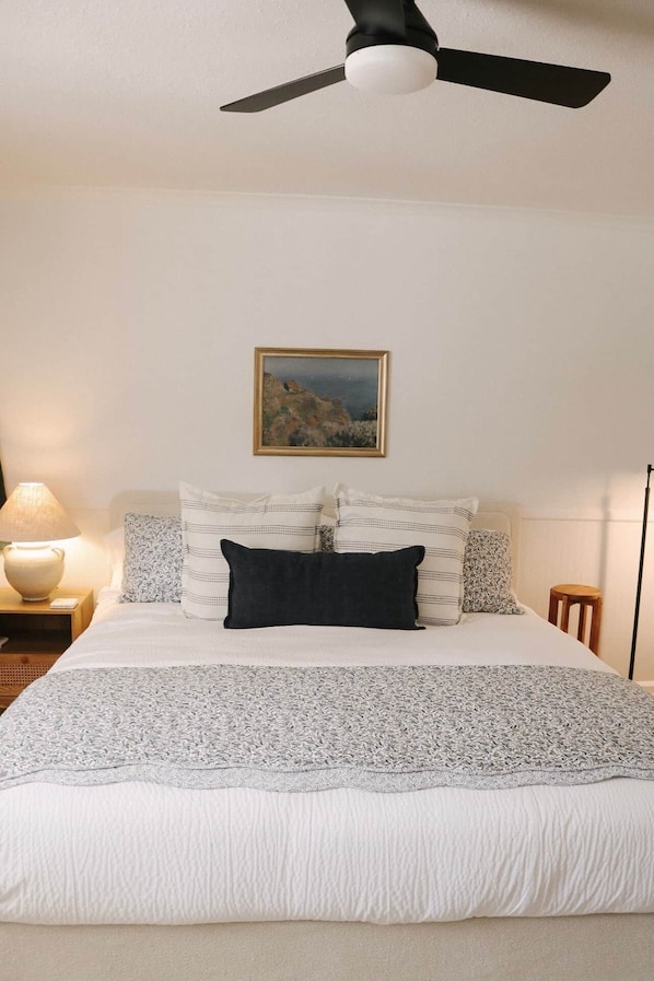 Ein Schlafzimmer mit Bett, Kissen, Nachttisch, Lampe, einem gerahmten Gemälde und Deckenventilator.