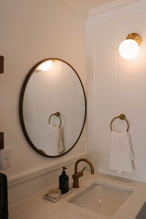 Een badkamer met een ronde spiegel, twee handdoekringen, een wastafel, een kraan en een wandlamp.