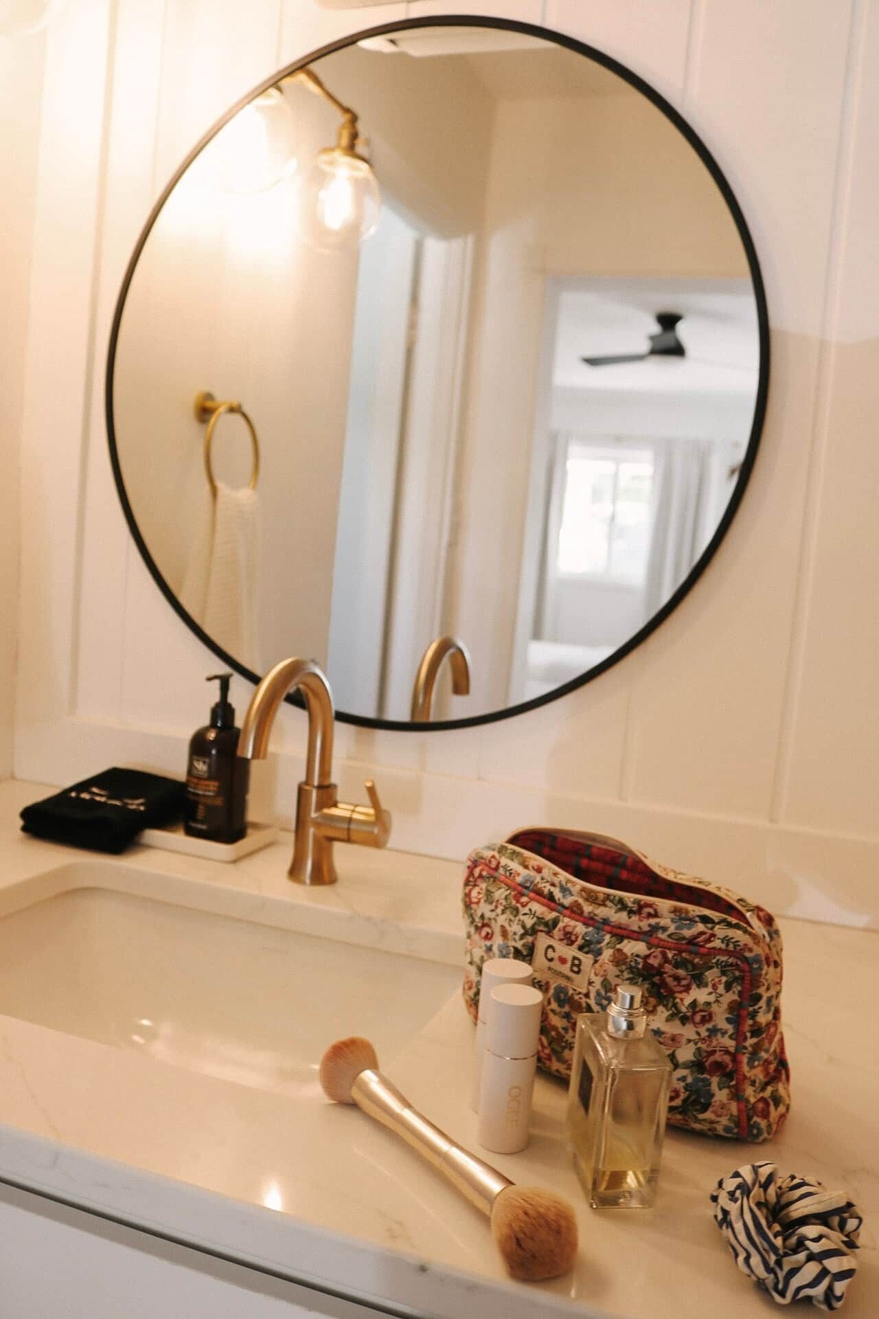 A bathroom with a round mirror, a gold faucet, a sink, and toiletries including a brush, a bottle, and a bag.