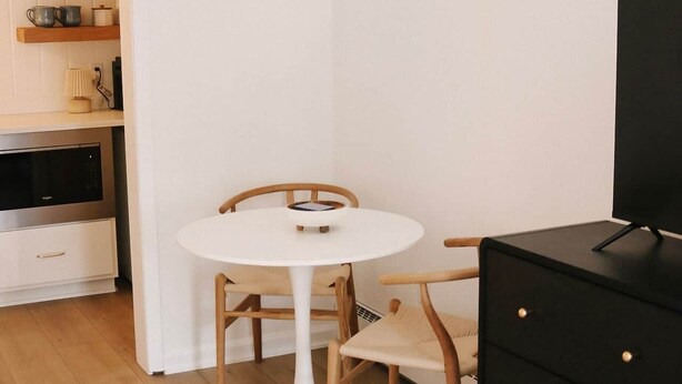 Ein gemütlicher, kompakter Wohnbereich mit einem runden weißen Tisch, Holzstühlen, einem schwarzen Schrank und einer Küchenzeile mit Mikrowelle und Regalen.