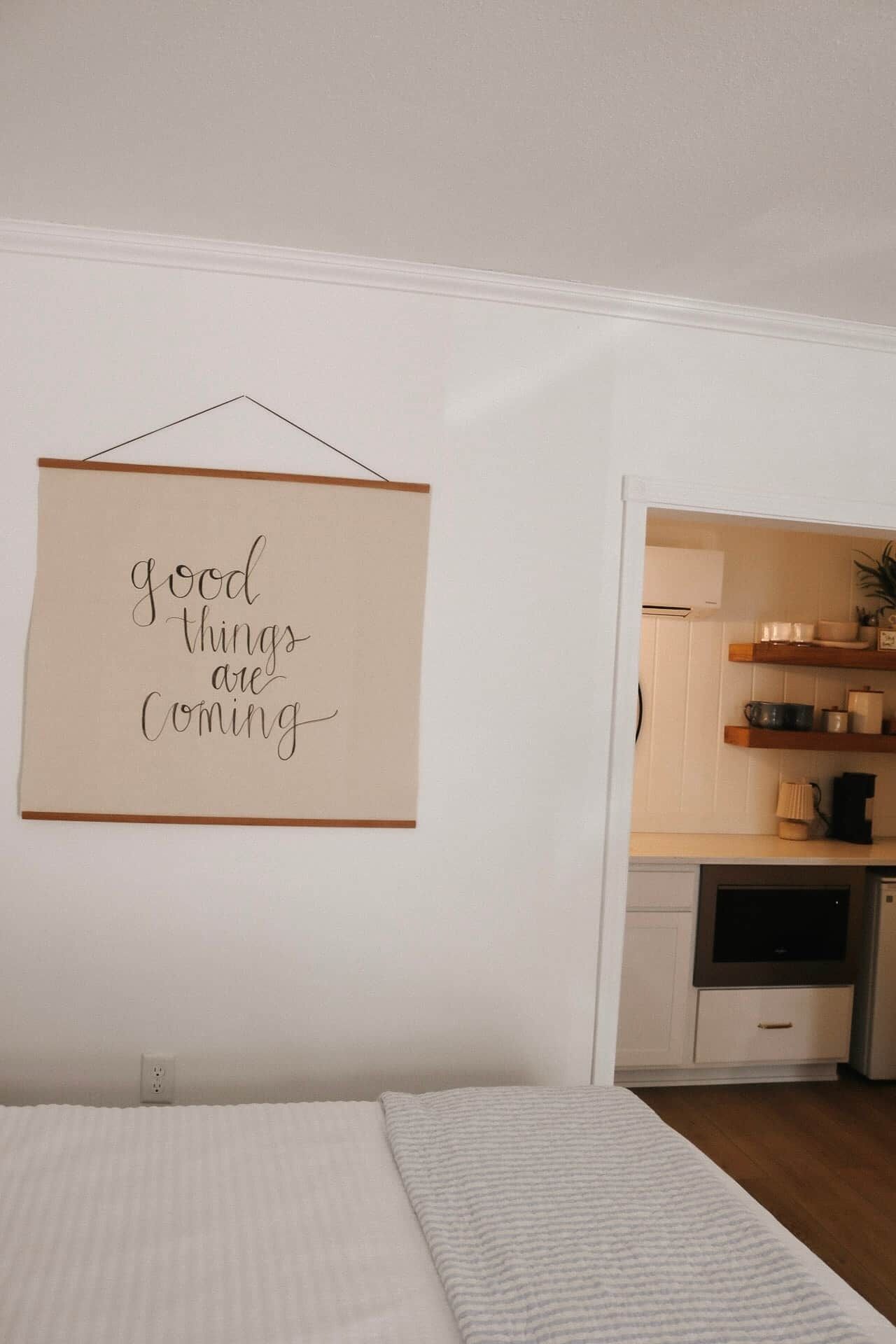A bedroom with a bed, a wall-mounted sign, a kitchen area with shelves, and a white door.