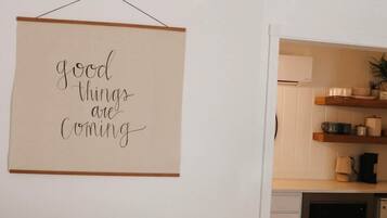 A bedroom with a bed, a wall-mounted sign, a kitchen area with shelves, and a white door.