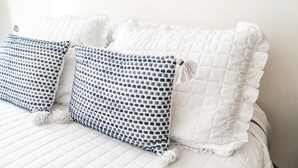 Un lit bien fait, avec une literie blanche et deux coussins à motifs, un tableau encadré au mur et une disposition simple et épurée de la pièce.