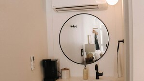 Une salle de bain avec un meuble-lavabo blanc, un miroir rond, une machine à café noire et un luminaire.