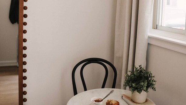 Un coin repas comprenant une table ronde, des chaises, un luminaire suspendu, un miroir et une fenêtre avec des rideaux.