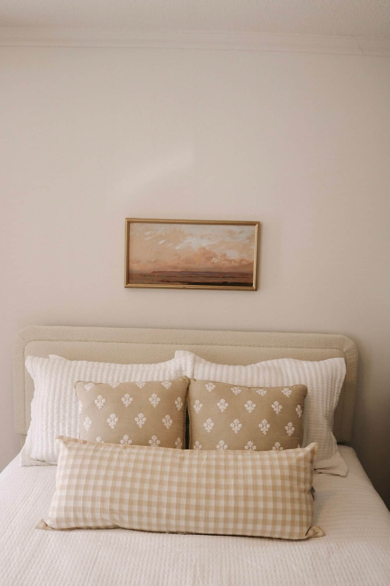 A bed with a beige headboard, white and beige patterned pillows, and a framed painting above.