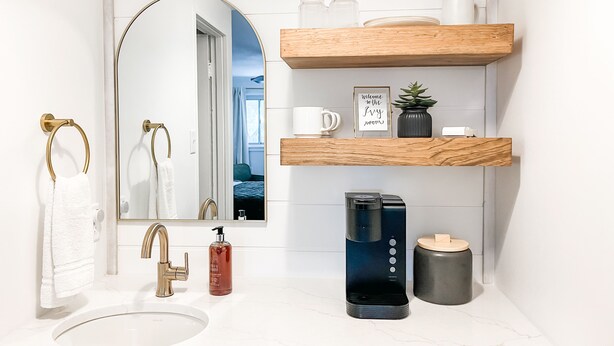 Un baño moderno con lavamanos blanco, detalles dorados, un espejo, una cafetera y estantes de madera con artículos de aseo.