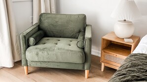 Una habitación con un sillón tapizado en verde, una mesita de noche de madera con una lámpara, y una cama con una colcha verde oscuro.
