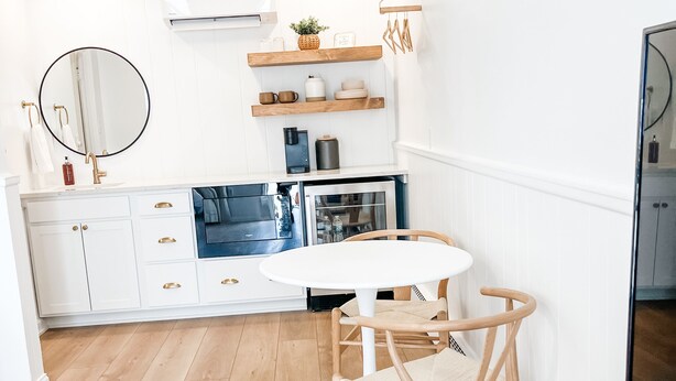 A modern kitchen with white cabinetry, a round mirror, wooden shelves, and a dining area with a white table and chairs. - The Goldie Room - #21 (Whitehall)