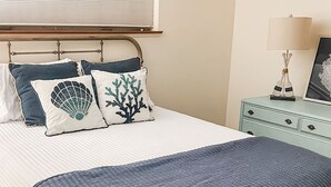 Een net opgemaakt bed met een wit-blauwe spreideken, twee sierkussens met zeemotieven, een blauwe plaid, een houten hoofdbord, een nachtkastje met een lamp en een klein dressoir.