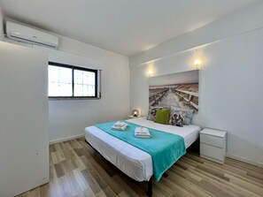 Un dormitorio con cama, mesita de noche, aire acondicionado de pared, ventana y un mural con motivos playeros.