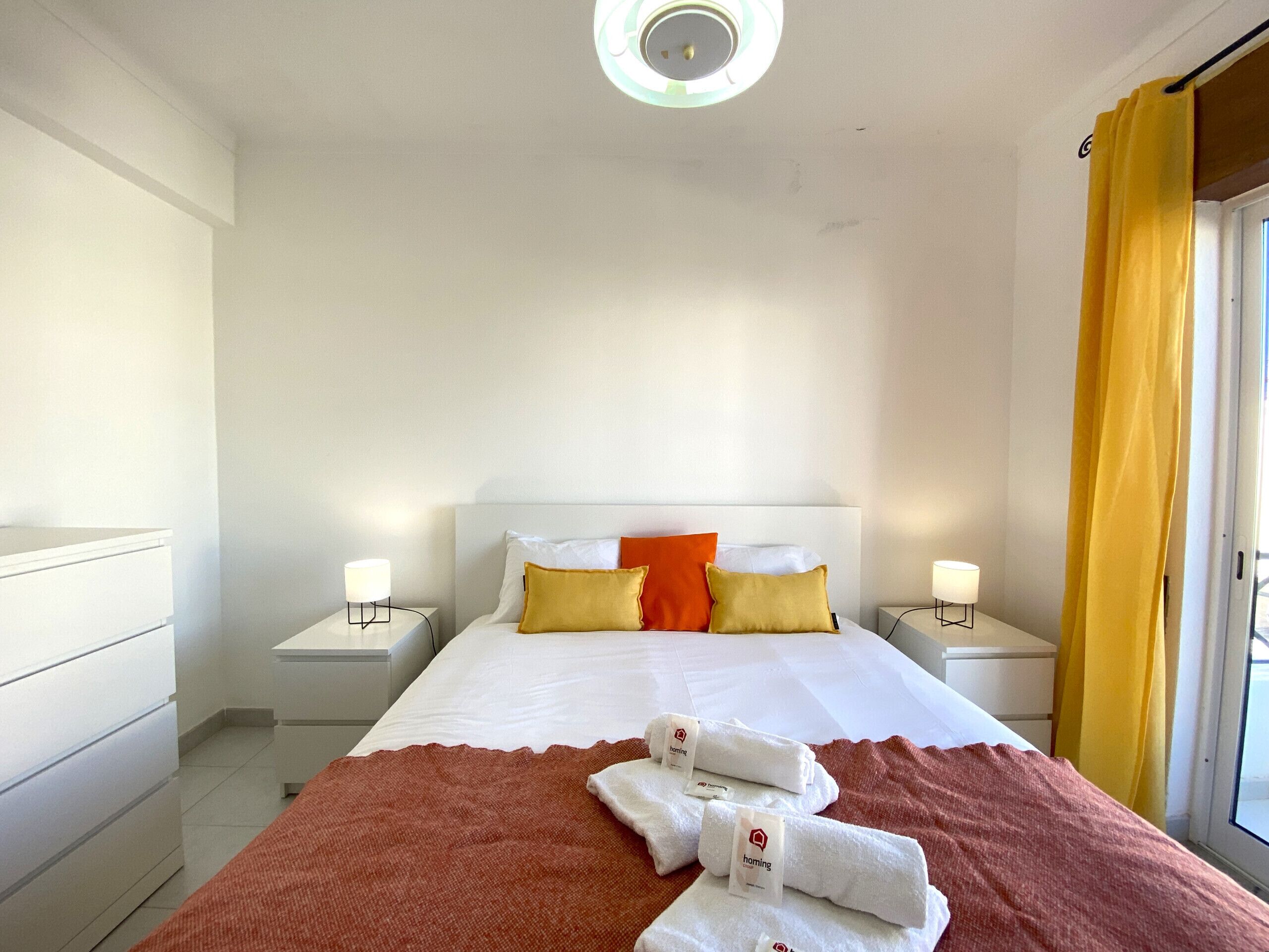 A hotel room with a bed, two bedside tables, a nightstand, a window with yellow curtains, and a ceiling light.