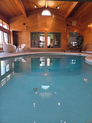 An indoor swimming pool with wooden walls and ceiling, surrounded by wicker furniture and a large arched window.