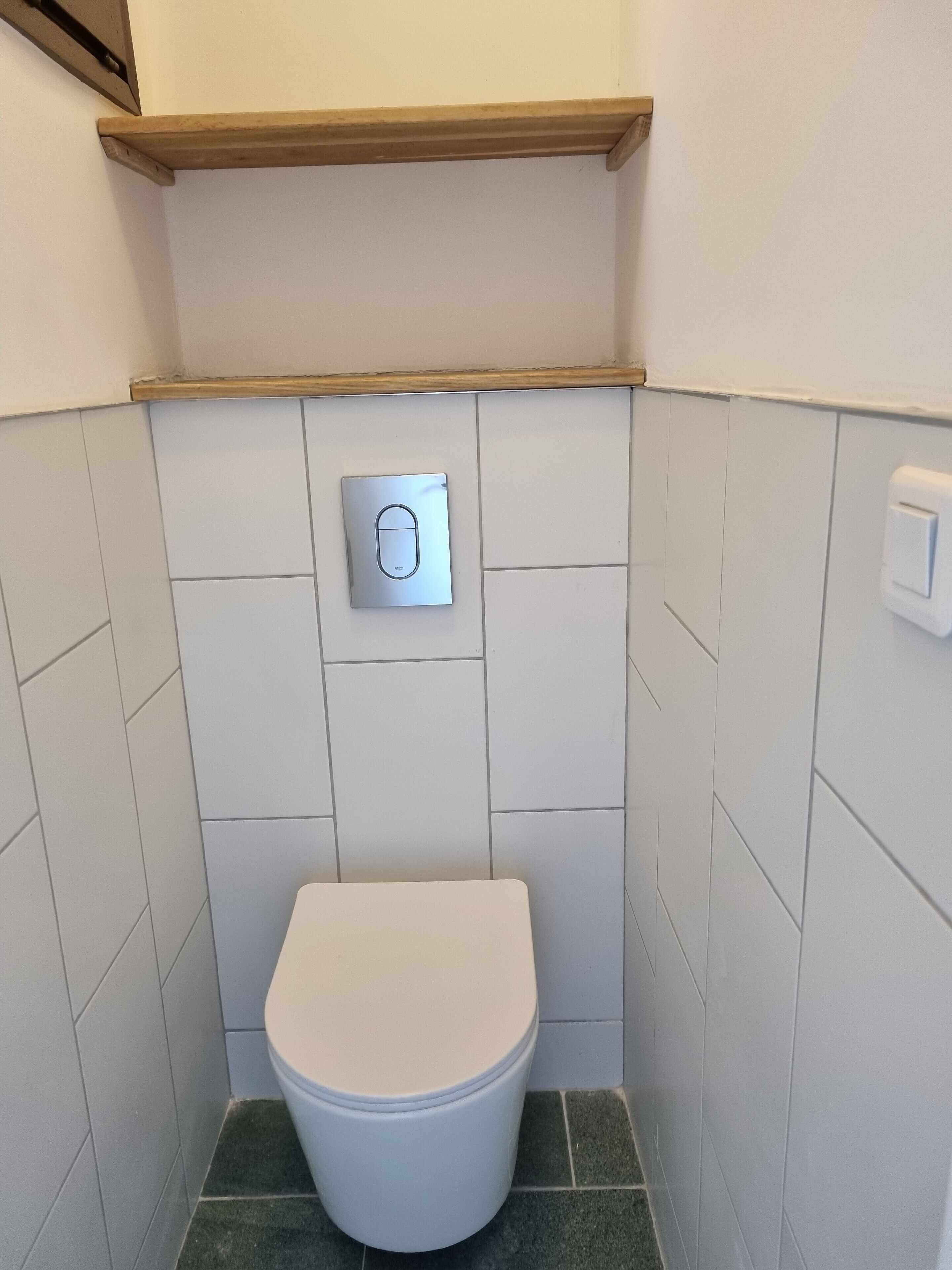 Um banheiro pequeno com azulejos brancos, vaso sanitário branco, alavanca de descarga e prateleira de madeira acima.