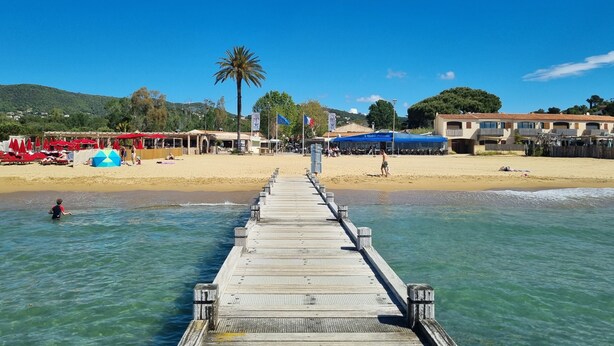 Ein Strand mit einer Holzsteg, der zum Ufer führt, einer Palme und Gebäuden mit Balkonen im Hintergrund.