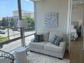 Una sala de estar moderna con un sofá gris, cojines estampados, una mesita hexagonal y un ventanal con vistas a la calle y edificios.