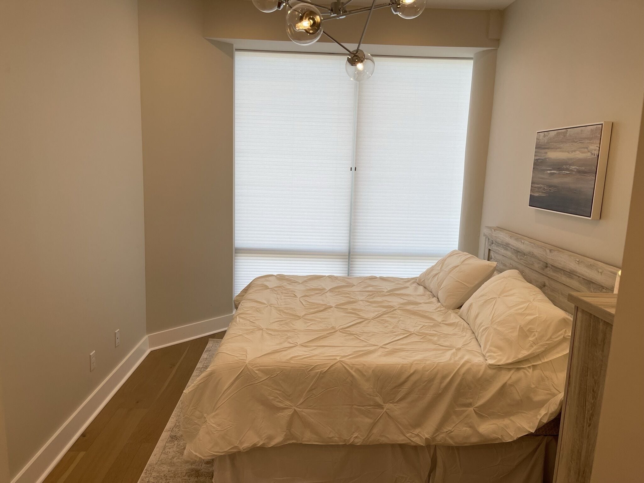 A bedroom with a bed, a headboard, a window with blinds, a ceiling light fixture, and a framed picture on the wall.