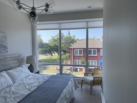 Un dormitorio con una cama grande, una silla y vistas a un edificio rojo a través de amplios ventanales.