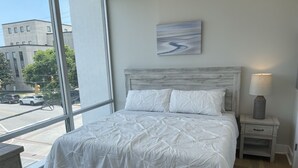 Quarto com uma grande janela, cama com roupa de cama branca, mesa de cabeceira com abajur e um quadro na parede.